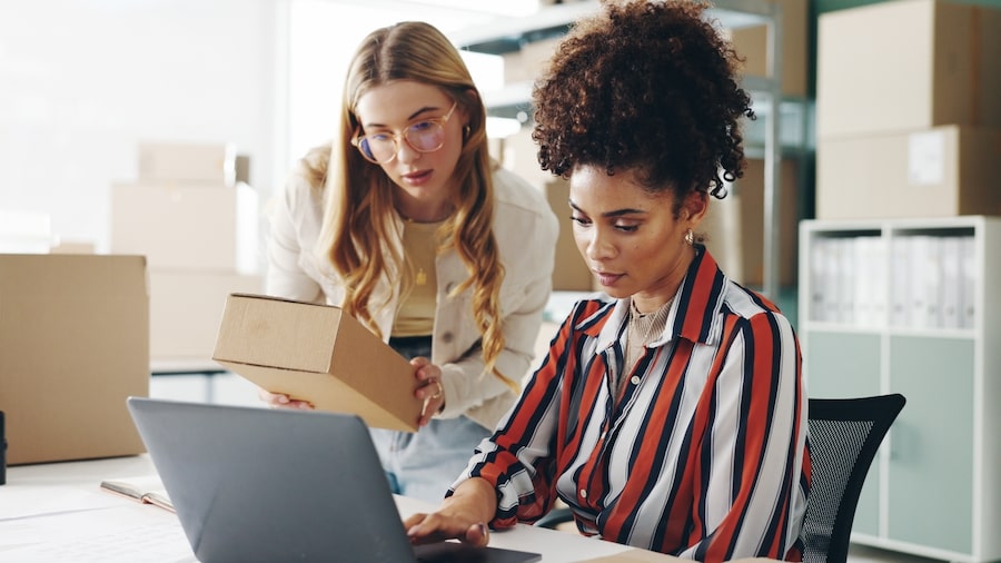 Two women looking at the computer’s screen, one holding a box.