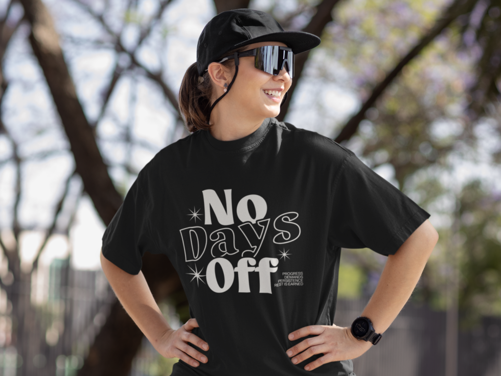 A woman with a ponytail wears a black cap and mirrored sunglasses, smiling confidently. Her black t-shirt reads "No Days Off".