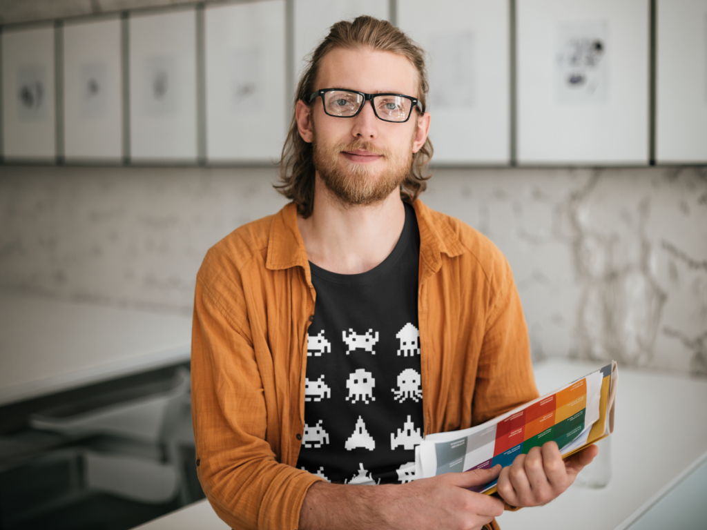 A man with glasses and a beard smiles while holding colorful charts. He wears a black shirt with white space invaders and an orange jacket.