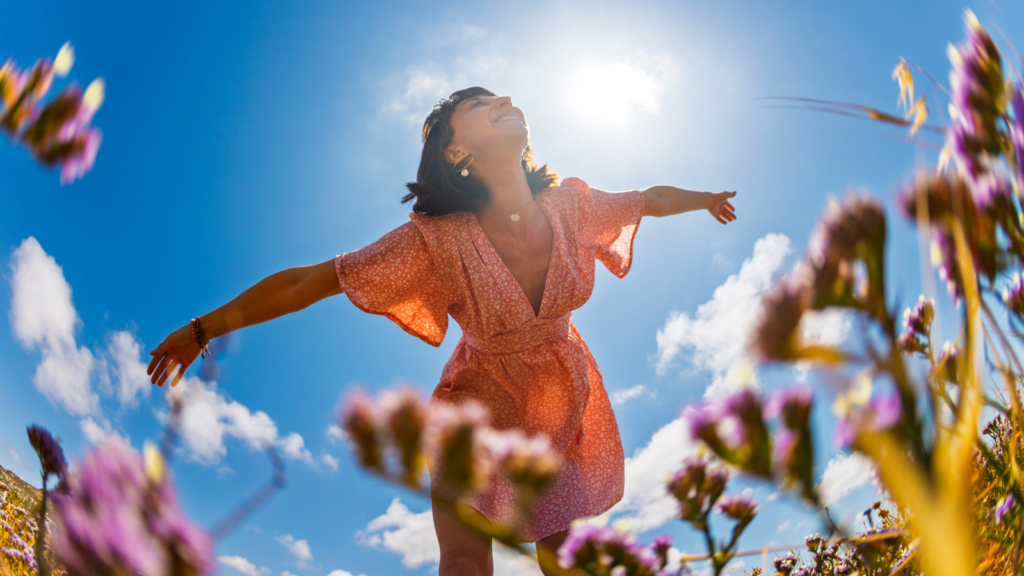 A joyful woman in a red floral dress spreads her arms under a bright sunlit sky, surrounded by purple flowers and a few clouds, conveying freedom and happiness.