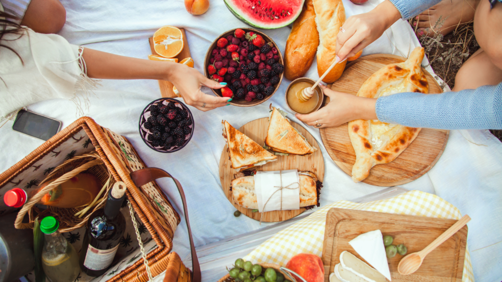 A vibrant picnic scene on a white blanket features sliced watermelon, berries, pastries, and a cheese platter.