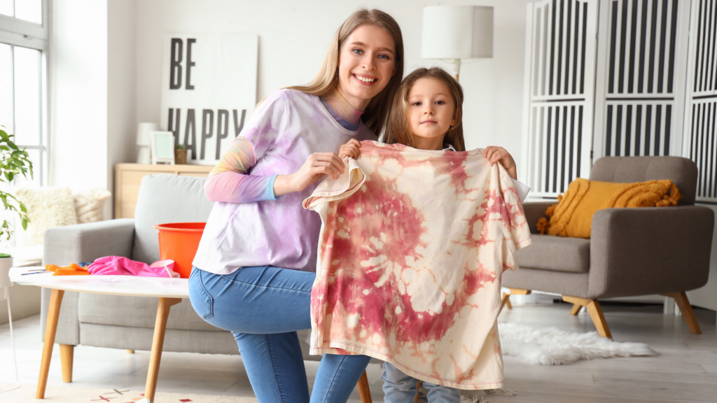 A woman and a child happily display a tie-dyed shirt in a cozy, bright living room with modern decor, promoting a joyful and creative atmosphere.