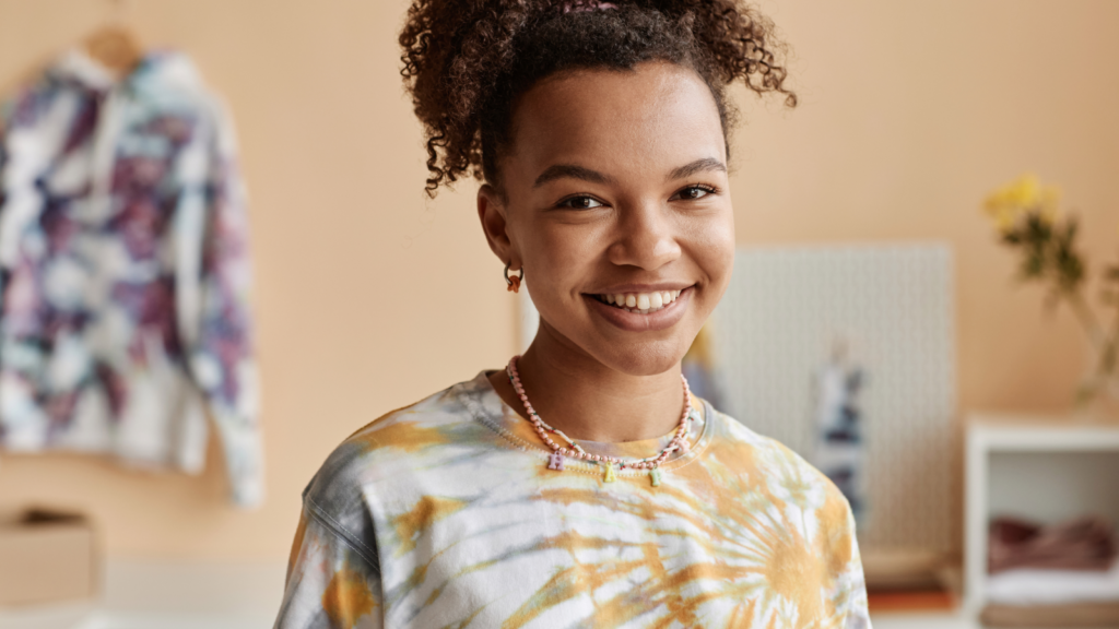 Smiling person in a tie-dye shirt and necklace stands in a cozy room.