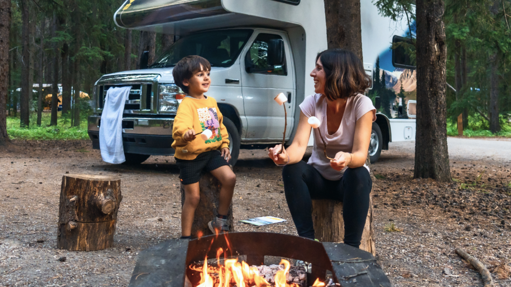 A woman and child toast marshmallows over a campfire in a forest, near an RV.