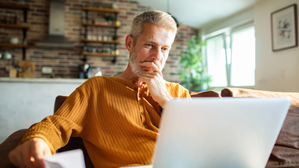 A middle-aged man in an orange sweater sits on a couch, thoughtfully looking at a laptop in a cozy, sunlit living room.