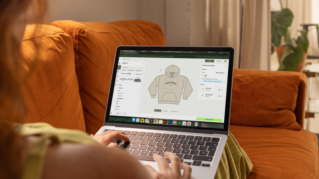 A woman sitting on an orange couch, using a laptop to design a beige hoodie with Printify’s Product Creator.