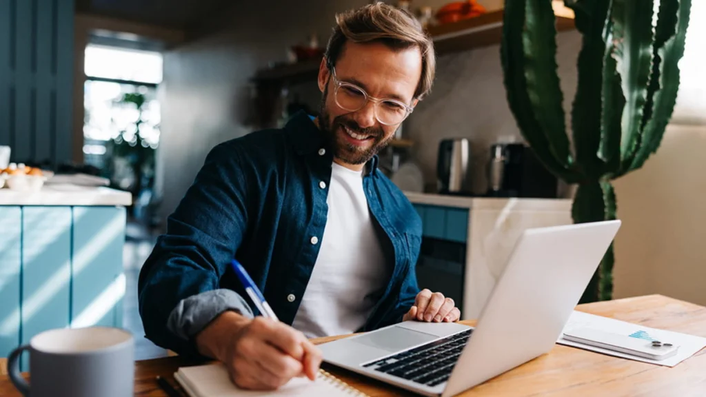 A smiling man with glasses writes in a notebook while using a laptop at a kitchen table. A large cactus is in the background.