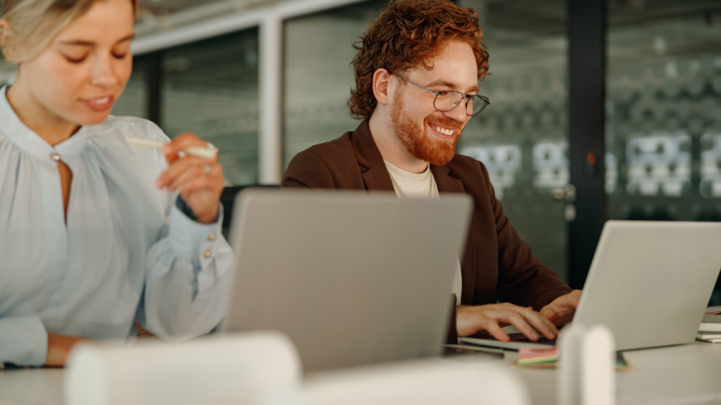 A man and woman sit together at a table, both working on laptops, immersed in their tasks.