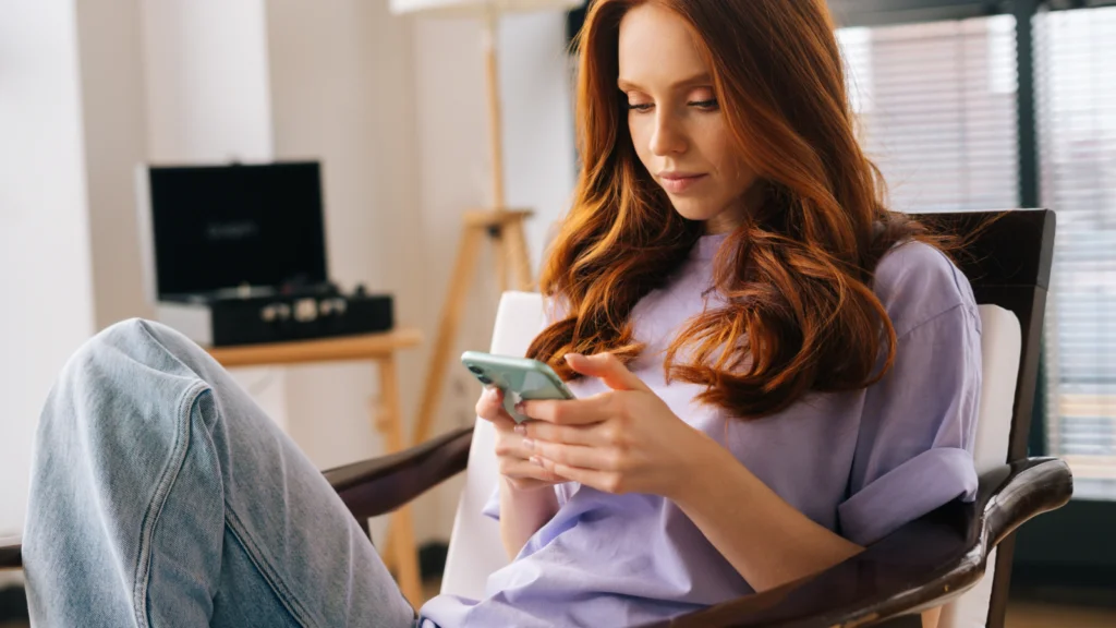 A young woman with long red hair sits comfortably in a chair, focused on her smartphone. She wears a lavender shirt and jeans.