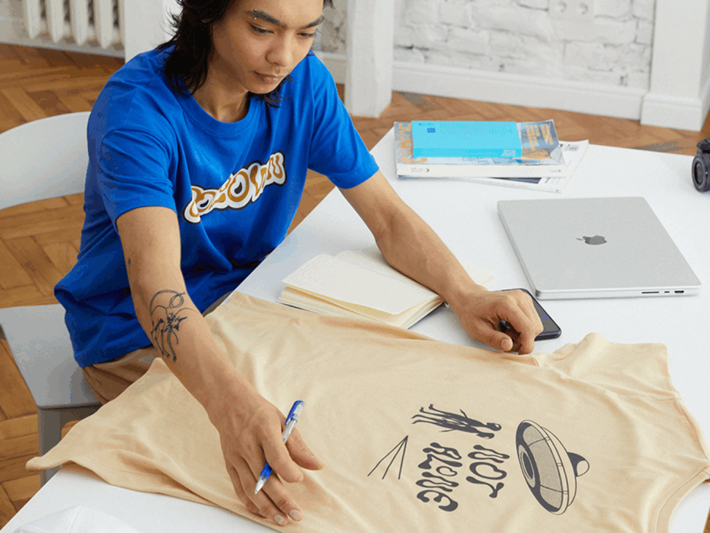 A young man in a blue t-shirt looking at a custom beige t-shirt.