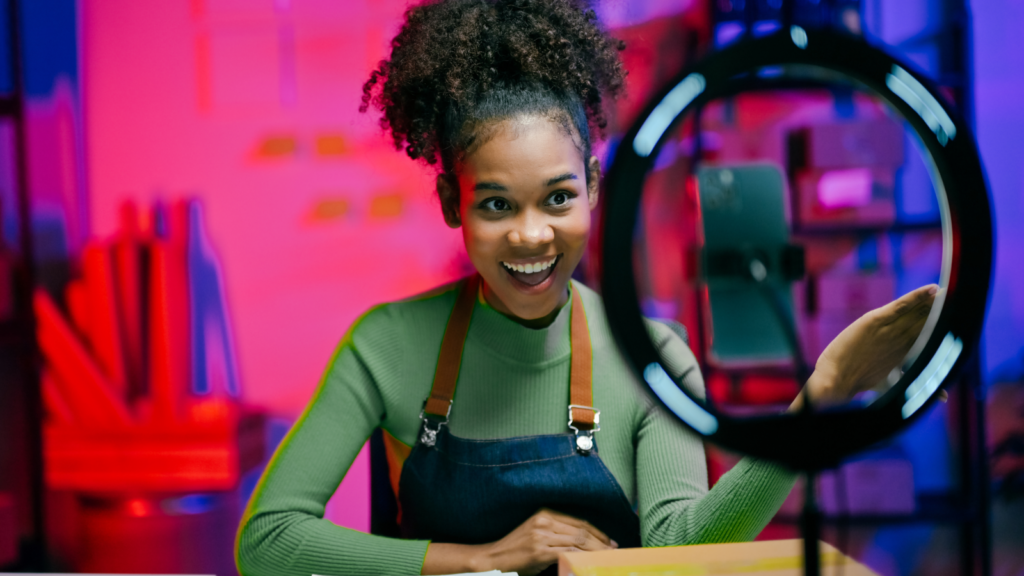 A smiling woman in a green top and apron, behind a ring light, recording on a smartphone.