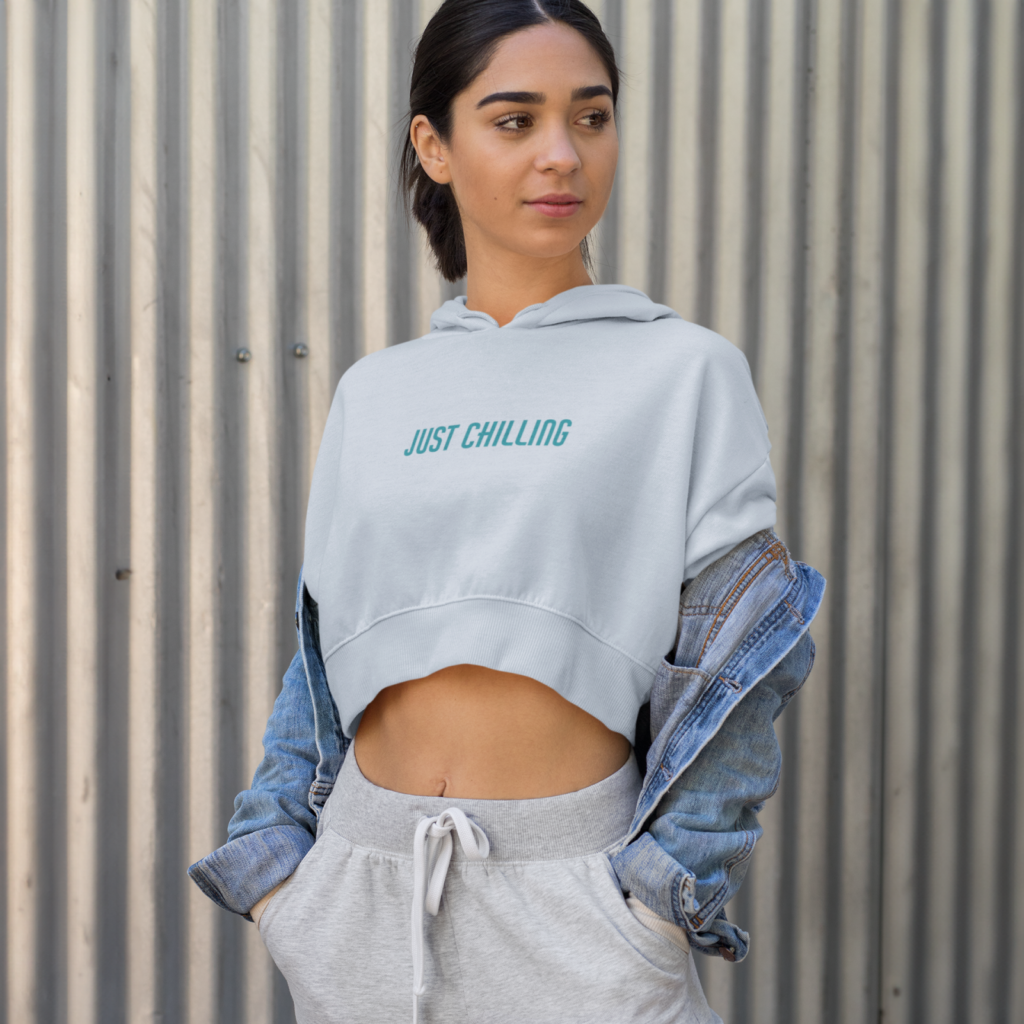 A person standing against a corrugated metal wall wearing a white hoodie that says "Just Chilling," a denim jacket, and gray sweatpants.