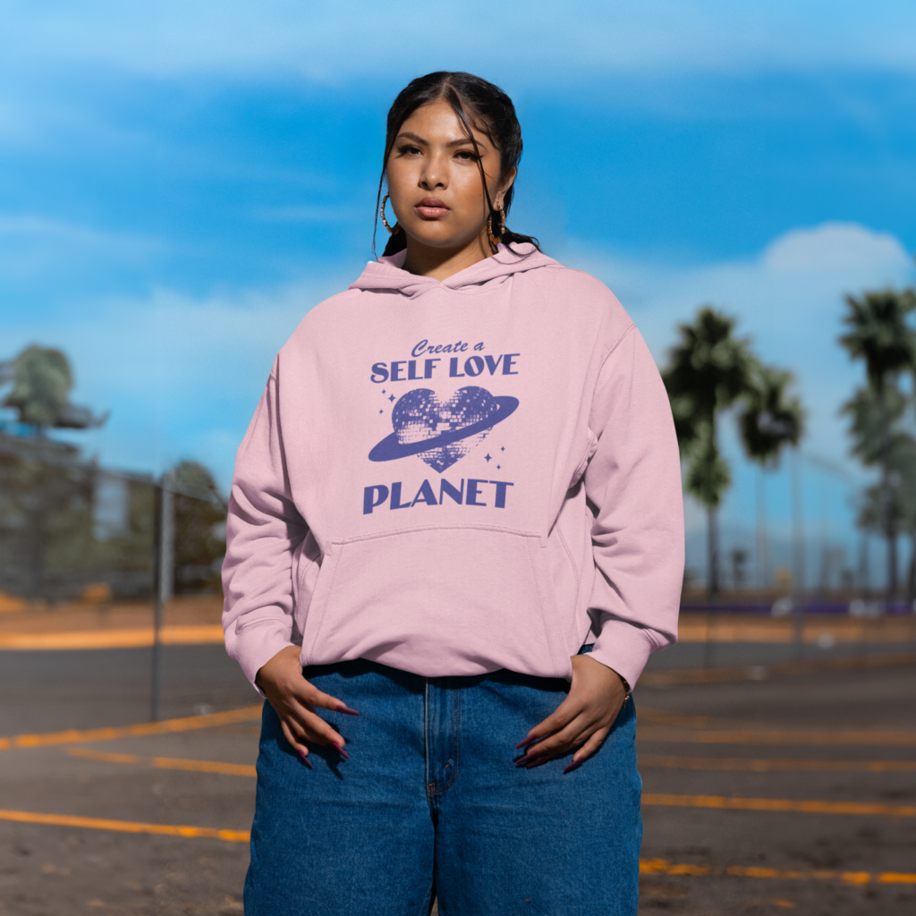 A woman in a pink “Create a self love planet” custom hoodie and blue jeans standing in a parking lot.