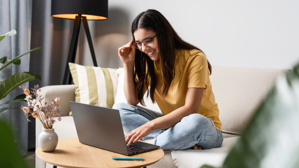 A woman wearing glasses sits cross-legged on a couch, smiling as she uses a laptop to learn how to create a Printify Pop-Up Store.