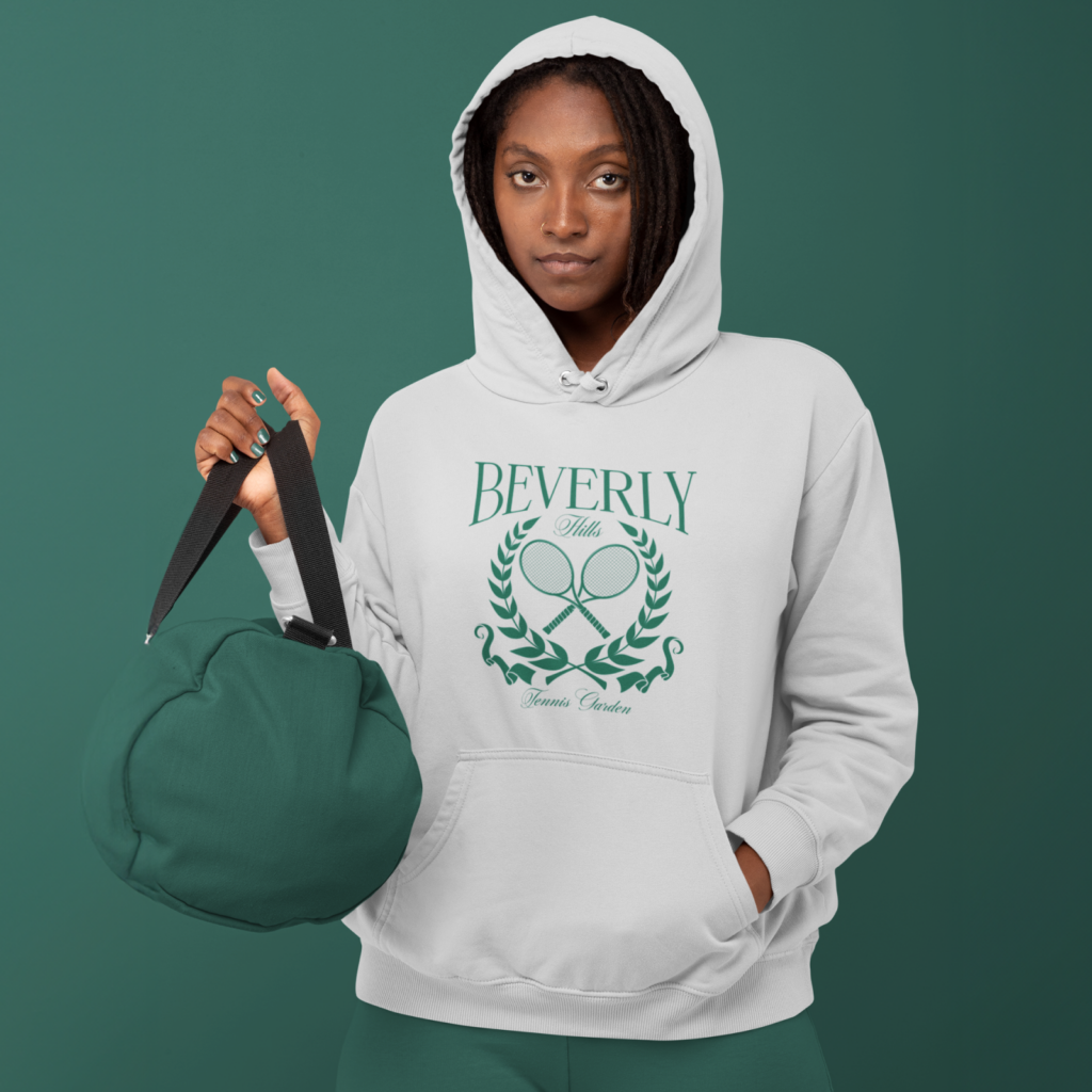 A woman in a white "Beverly Hills Tennis Club" hoodie holding a green bag against a green background.